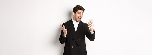 Free photo portrait of happy smiling man in black suit looking at smartphone screen with relieved and cheerful