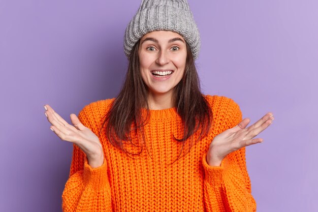 Portrait of happy smiling woman raises palms makes clueless gesture shrugs shoulders wears winter orange jumper and hat has dark hair.