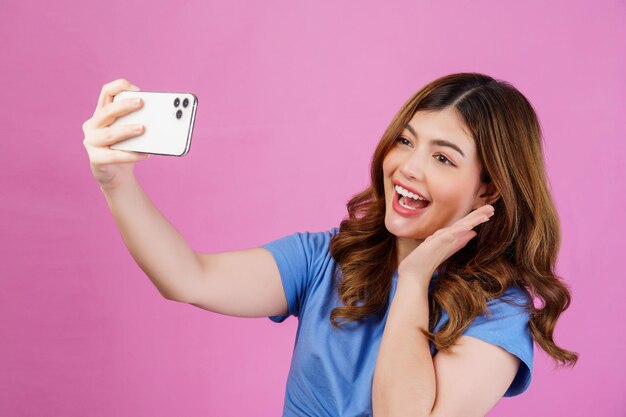 Portrait of happy smiling young woman wearing casual tshirt selfie with smartphone isolated over pink background