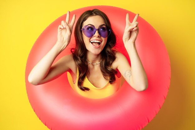 Portrait of happy stylish girl in sunglasses, inside beach swimming ring, showing peace sign, laughing and smiling, standing over yellow background