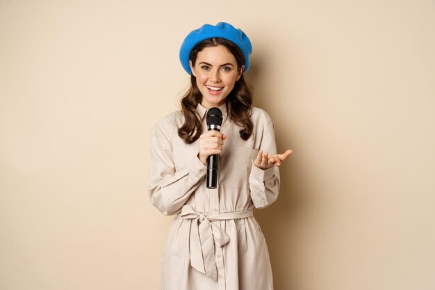 Portrait of happy stylish woman performing singing with microphone posing against beige background