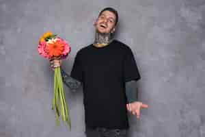 Free photo portrait of a happy tattooed young man holding gerbera flowers in hand shrugging against grey background