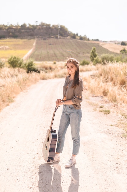 Portrait of a happy teenage girl with guitar standing on dirt track