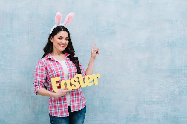 Free photo portrait of a happy woman with bunny ears on head holding easter word pointing finger upward on blue background