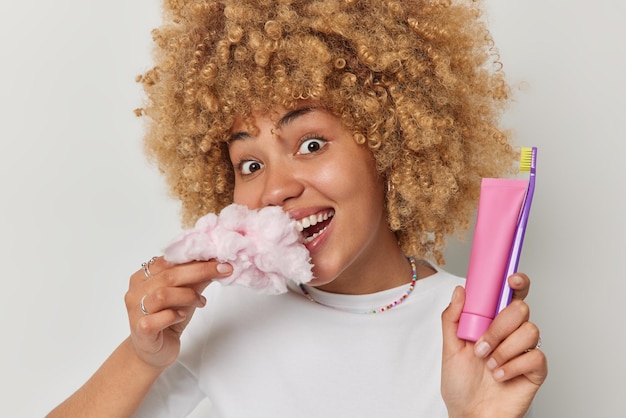 Free photo portrait of happy woman with curly hair eats cotton candy harmful for your teeth holds tube of toothpaste and toothbrush has glad expression poses against white background oral hygiene and sweet food