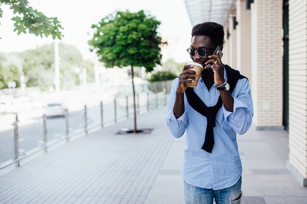 Portrait of happy young african man talking on phone and walking on the street with cup of coffee.