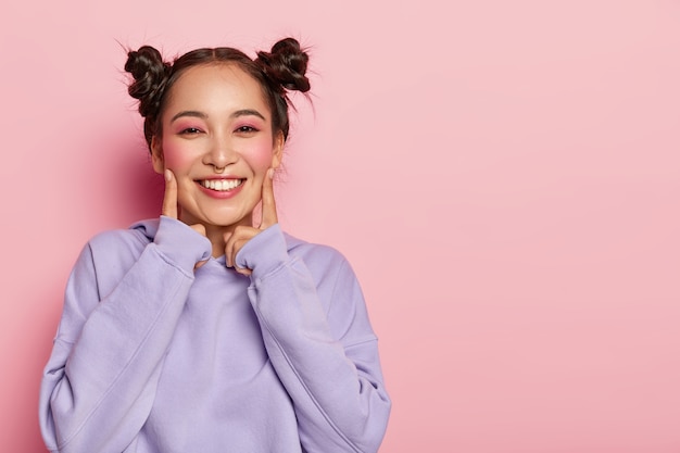 Portrait of happy young Asian girl stands indoor, touches cheeks with index fingers , has pleasant smile on face, dressed in casual purple hoody, wears pinup makeup