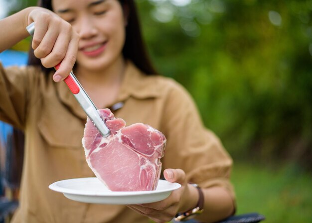 Portrait of happy young asian woman camping alone grilled pork barbecue in the picnic pan and cooking food while sitting on chair in the camping site