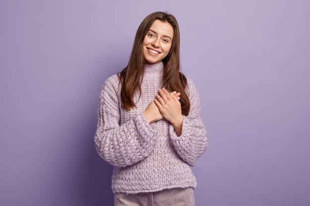 Free photo portrait of happy young european woman keeps hands on breast, shows heart gesture, expresses gratitude, being thankful, models against purple wall body language. monochrome. people and devotion