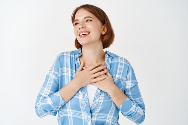 Portrait of happy young girl holding hands on heart smiling grateful say thank you express gratitude and fascination looking aside at copy space white background