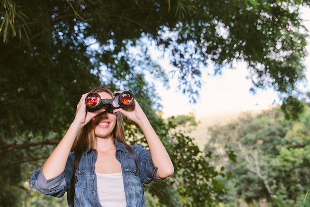 Portrait of a happy young woman looking through binoculars in forest