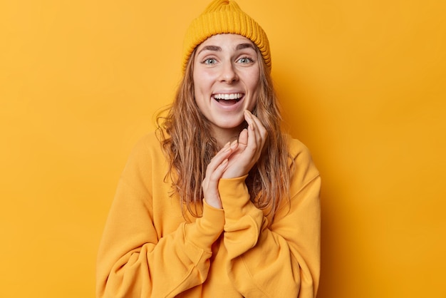 Portrait of happy young woman smiled toothily has surprised expression perfect white teeth laughs joyfully reacts on awesome news wears casual sweatshirt and hat isolated over yellow background