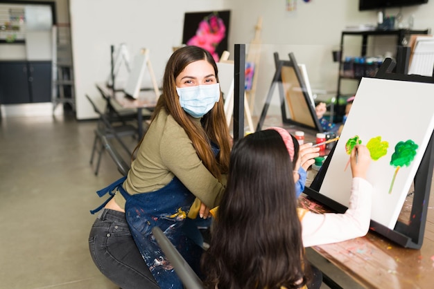 Portrait of a hispanic female teacher with a face mask teaching a girl child how to paint with a brush during a painting class