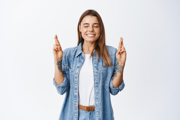 Portrait of hopeful dreamy girl close eyes and making a wish cross fingers for good luck and praying believe in dreams come true imaging something positive white background