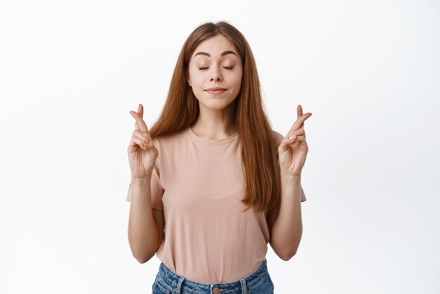 Portrait of hopeful girl student wishing about exam cross fingers for good luck and close eyes praying pleading for dream come true white background