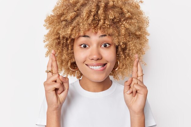 Portrait of hopeful woman anticipates for important results bites lips keeps fingers crossed makes wish has happy expression wears basic t shirt poses against white background. Body language