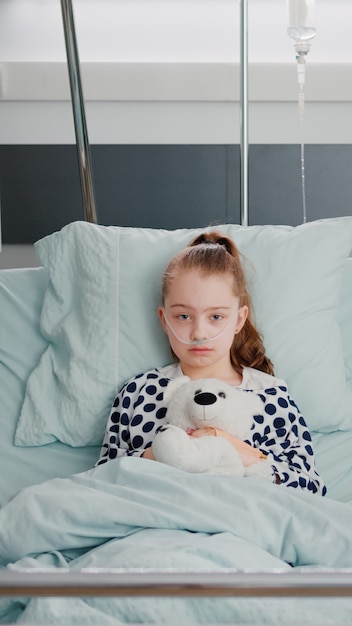 Free photo portrait of hospitalized sick girl child patient holding teddy bear resting in bed during medical co...