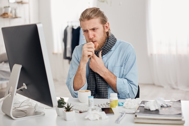 Portrait of ill sick bearded male manager coughs, has cold and flu. Young fair-haired man has running nose, cough and bad cold, sits at workplace in front of computer screen. Illness and infection