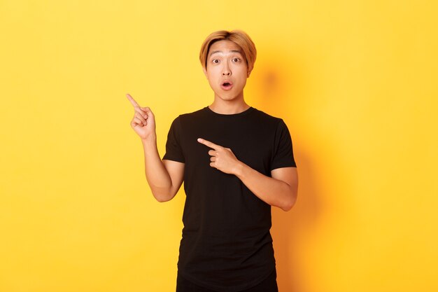 Portrait of impressed and excited handsome asian guy in black t-shirt, react to your logo, pointing fingers upper left corner