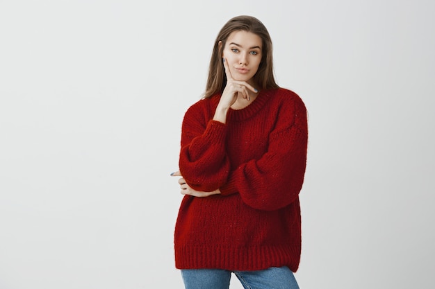 Portrait of indifferent unimpressed attractive feminine european female in stylish loose sweater, holding fingers on cheek and looking with displeased expression, showing contempt 