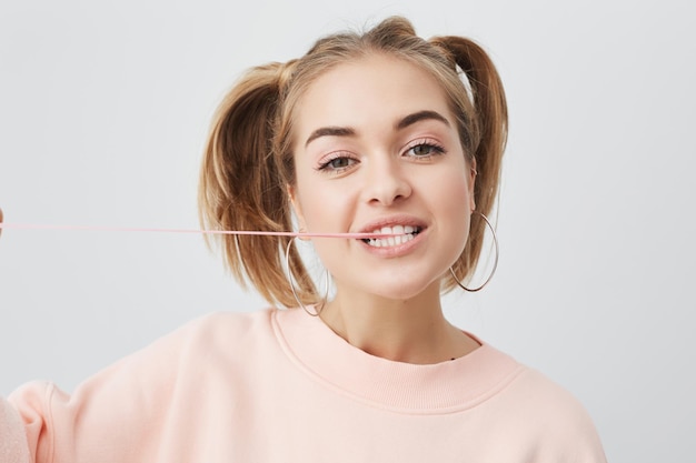 Portrait of joyful cheerful teenage girl with healthy clean skin dark eyes two ponytails in pink sweater posing against gray background showing her white teeth and stretching chewing gum