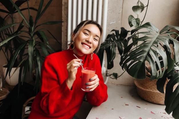 Free Photo portrait of joyful lady in red oversized sweater holding glass of freshly squeezed watermelon juice, sitting in cafe against wall of plants