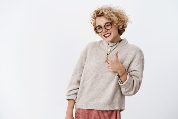 Portrait of joyful satisfied and impressed good-looking charming cheerful fair-haired female in glasses and sweater showing thumb up and winking flirty, liking and agree to awesome plan.