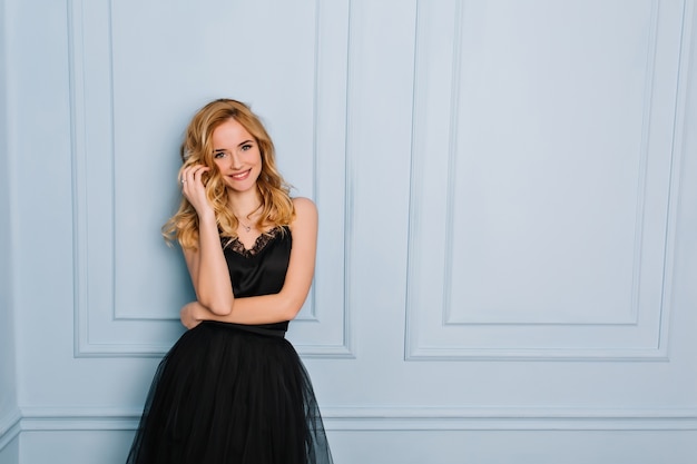 Free Photo portrait of joyful young blonde woman wearing elegant black lace dress. blue wall. her mouth opened, touching her face. she has long blonde wavy hair.