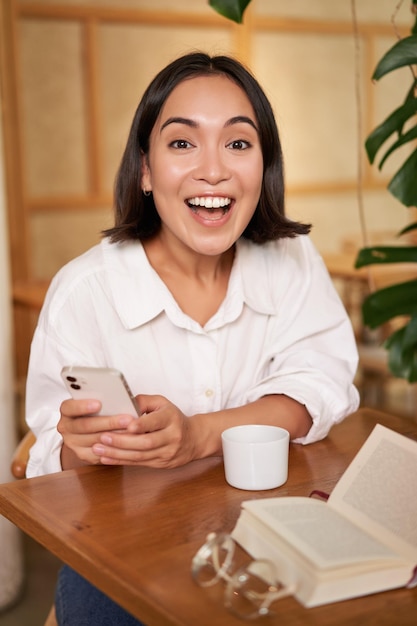 Free photo portrait of korean girl in cafe drinking coffee using mobile phone and reading a book in cozy place