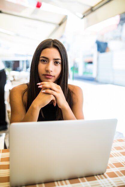 Portrait of latin woman in glasses working with her laptop outdoors. Charming freelancer woman with curly hair is sitting in street bar and doing her remote work on the netbook