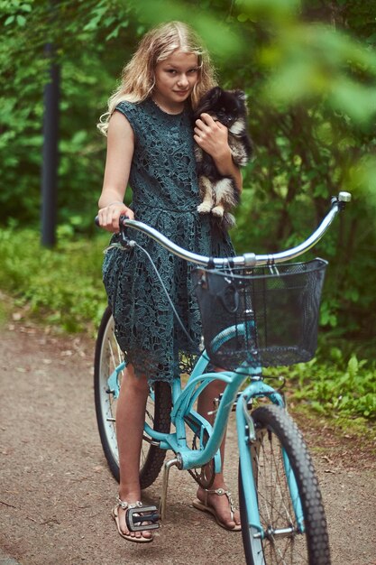 Portrait of a little blonde girl in a casual dress, holds cute spitz dog. Ride on a bicycle in a park.