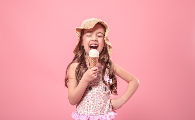 Free photo portrait of a little cheerful girl in a summer hat with ice cream in her hands, on a colored pink background, summer concept