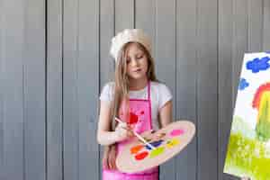 Free photo portrait of a little girl standing against grey wall painting with brush against gray wall