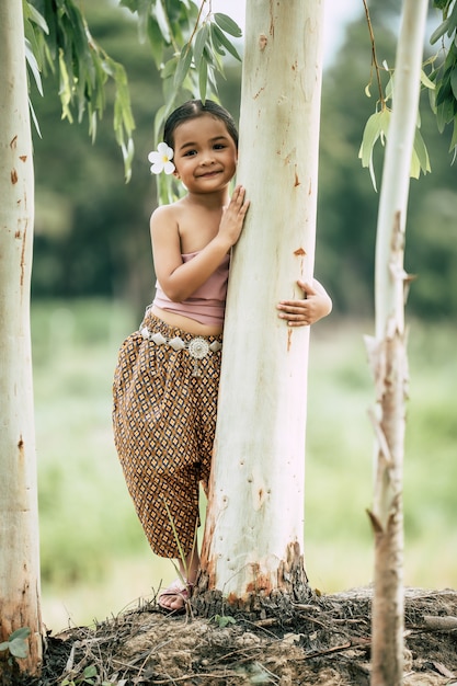 Free photo portrait of little girl  in thai traditional dress and put white flower on her ear, stand and embrace trunk of the tree, smile , copy space