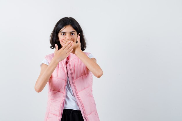 Portrait of a little girl in white t-shirt and puffer vest covering her mouth