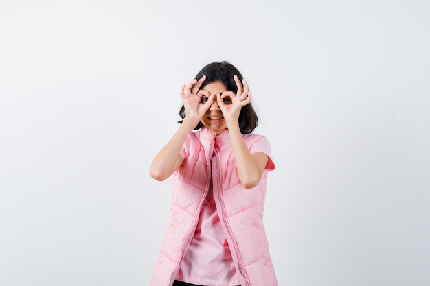 Portrait of a little girl in white t-shirt and puffer vest showing glasses gesture