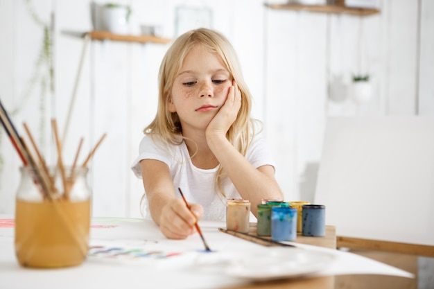 Portrait of little girlwith blonde hair and freckles sitting at the desk and, putting her elbow on the table