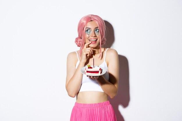 Portrait of lovely birthday girl in pink party wig, holding cake, thinking before making wish, standing over white background