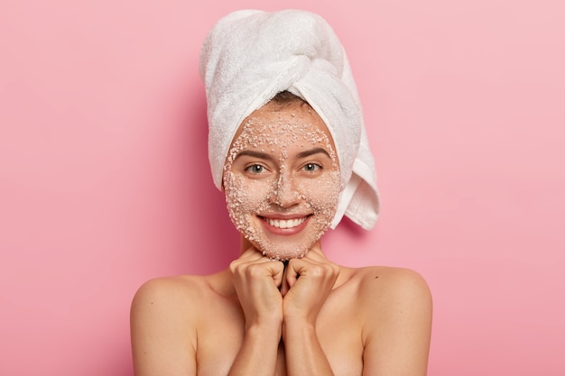 Portrait of lovely cheerful woman smiles gently, enjoys softness of skin after beauty procedures, smiles pleasantly, shows white teeth, has adorable look at camera, poses against pink background