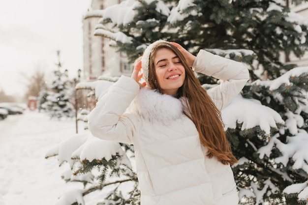 Portrait lovely cute woman chilling on sunshine in frozen morning. Young joyful woman enjoying winter time on fir trees full with snow. Positive true emotions, smiling with closed eyes.