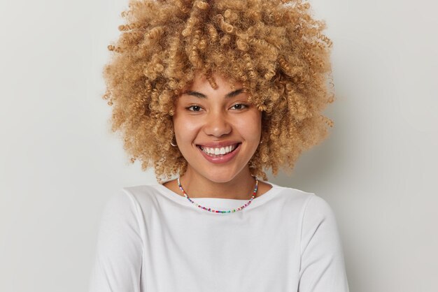 Portrait of lovely happy curly haired woman smiles toothily looks with glad expression wears casual t shirt and necklace stays positive isolated over white background People and emotions concept