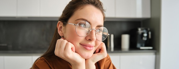 Free Photo portrait of lovely happy young woman wearing glasses sitting in kitchen with dreamy thoughtful face