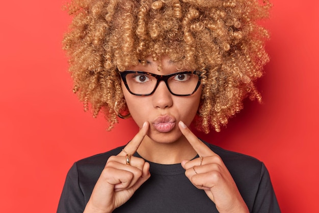 Free Photo portrait of lovely woman with curly blonde hair keeps lips folded indicates index fingers at mouth wears transparent glasses and casual black t shirt poses against red background sends air kiss