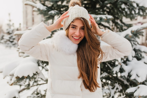 Free Photo portrait of  lovely woman with long light-brown hair showing true happy emotions in winter day on fir tree. charming young woman in white jacket fooling around in cold morning at snowy park.