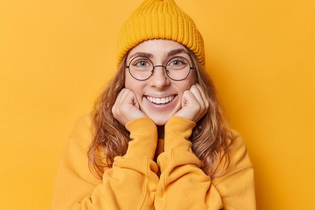 Portrait of lovely young European woman keeps hands under chin smiles broadly shows white teeth focused directly at camera wears casual sweatshirt and hat isolated over yellow studio background