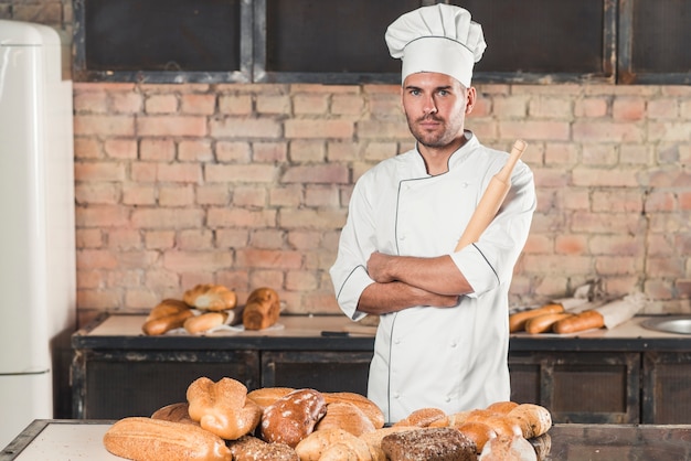 Free photo portrait of male baker holding rolling pin standing behind the baked breads on table