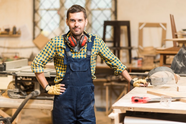 Free photo portrait of a male carpenter with his hand on hip standing near the workbench