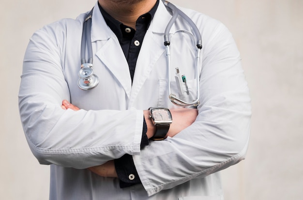 Portrait of a male doctor holding stethoscope around her neck standing with arms crossed against white backdrop