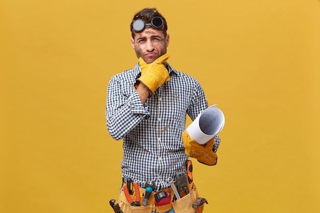 Free photo portrait of male fettler wearing protective wear holding blueprint having thoughtful expression while thinking over his actions during work. repairman with kit of instruments isolated over wall