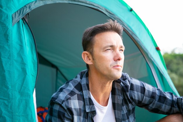 Portrait of male tourist sitting in tent and looking away. Young Caucasian hikers or traveler relaxing on nature and enjoying scenery. Backpacking tourism, adventure and summer vacation concept